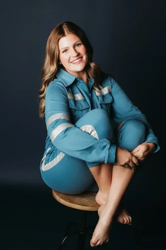 امرأة شابة مبتسمة ترتدي بدلة زرقاء، تجلس بشكل مريح على كرسي خشبي، مع خلفية داكنة تضفي طابعاً عصريًا.