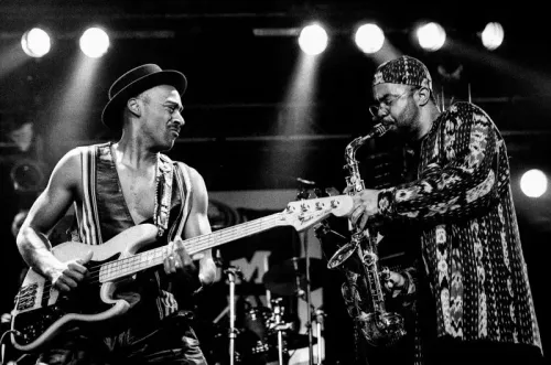 عازف الجيتار ماركوس ميلر يعزف في عرض حي، بينما يعزف عازف الساكسفون بجانبه، وسط أضواء المسرح.