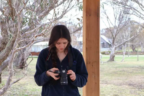 طالبة ترتدي سترة زرقاء، تحمل كاميرا، وتفحص الإعدادات، بينما تتواجد في منطقة طبيعية محاطة بالأشجار، في سياق حديث عن حظر وسائل التواصل الاجتماعي في أستراليا.