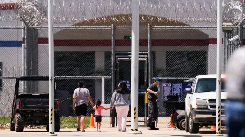 امرأتان تحملان أطفالًا صغيرين، تسيران نحو مركز احتجاز محاط بسياج شائك، مما يعكس قضايا الترحيل العائلي في الولايات المتحدة.