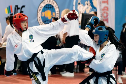 صورة لمنافسة في التايكوندو تُظهر لاعبتين تتبادلان الضربات في بطولة، مع التركيز على تقنيات الدفاع والهجوم.