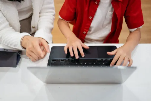 طفل يرتدي قميصًا أحمر يجلس أمام كمبيوتر محمول، بينما تساعده يد شخص بالغ في استخدامه، مما يعكس اهتمام الآباء بفتح حسابات ترامب للأطفال.