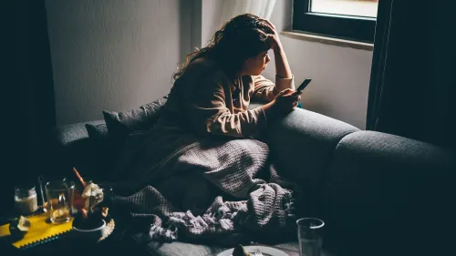 امرأة جالسة على أريكة، تمسك بهاتفها، تبدو مشغولة بالتفكير، محاطة بأشياء تشير إلى العزلة والتأمل في تأثير وسائل التواصل الاجتماعي على حياتها.