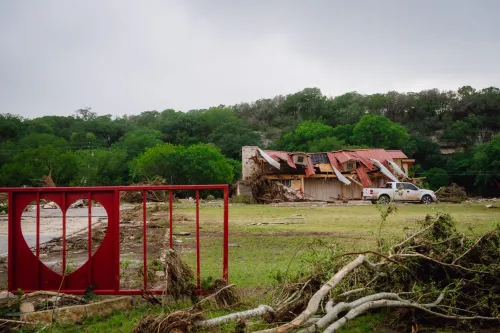 منزل متضرر وجدار حديدي أحمر عليه شكل قلب وسط آثار فيضان نهر Guadalupe في Texas Hill Country مع وجود شاحنة بيضاء.
