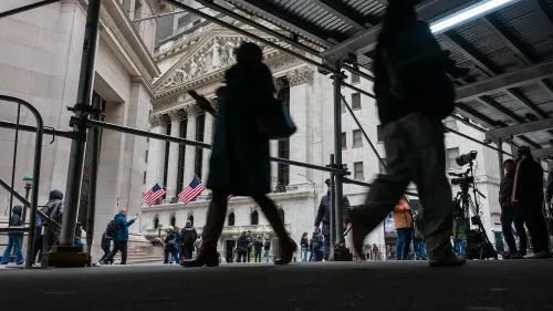 مشهد من أمام بورصة نيويورك، حيث يسير الناس تحت هيكل مؤقت، مع أعلام أمريكا ترفرف في الخلفية، مما يعكس حالة السوق المالية.