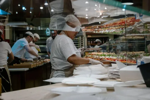 موظف في مطعم يقوم بتحضير الطعام، مع وجود زملاء في الخلفية، ويظهر تنوع الأطباق. الصورة تعكس نشاط قطاع الضيافة في سوق العمل.