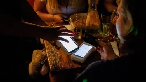 تظهر الصورة شخصين في مطعم يتبادلان الهواتف وبطاقة ائتمان، مما يبرز تأثير قانون ترامب الضريبي على الإكراميات.