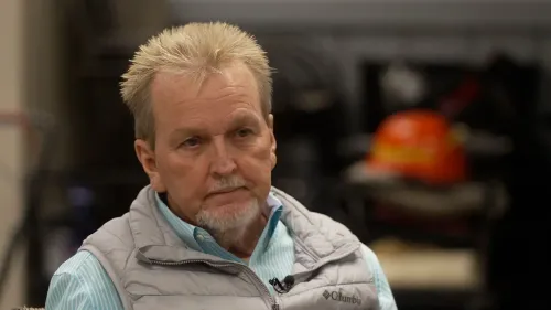 رجل مسن ذو لحية قصيرة يجلس في مكان داخلي، يبدو عليه التفكير العميق، مع أدوات خلفه تشير إلى بيئة عمل.