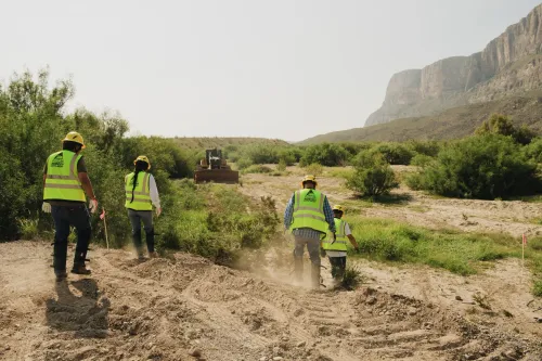 عمال يرتدون سترات أمان صفراء يعملون على مشروع تأمين الحدود في حديقة Big Bend الوطنية بتكساس وسط تضاريس جبلية ونباتات صحراوية.