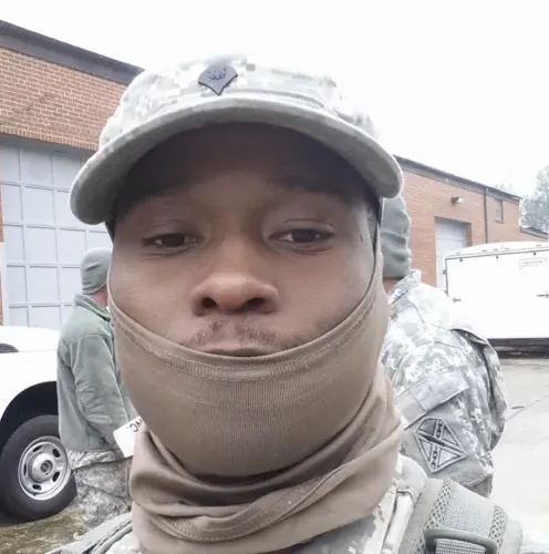 صورة لمسلح سابق في الحرس الوطني الأمريكي، محمد بيلور جالوه، يرتدي زيًا عسكريًا وقبعة، مع خلفية تظهر أشخاصًا آخرين في ملابس عسكرية.