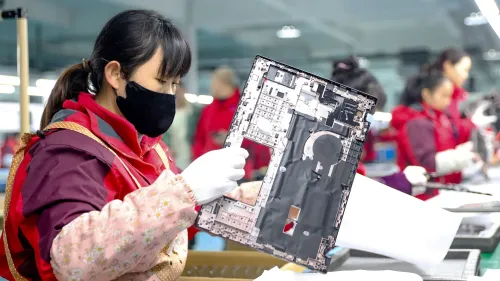 عاملة في مصنع تجميع الإلكترونيات تضع مكونات جهاز كمبيوتر، مع وجود عاملات أخريات في الخلفية، مما يعكس تأثير التعريفات الجمركية على الإلكترونيات.