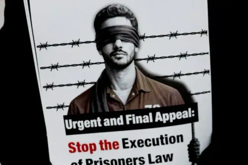 رجل معصوب العينين يرتدي زي سجن، يحمل لافتة تطالب بوقف قانون الإعدام، وسط سياج شائك، في سياق الاحتجاجات ضد التشريع الإسرائيلي.