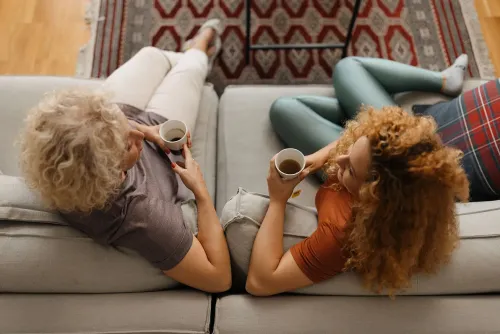 امرأتان تجلسان على أريكة، تتناولان مشروبات ساخنة، وتتناقشان في جو مريح. تعكس الصورة الروابط الأسرية وتبادل الدعم المالي.