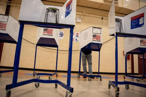 مركز اقتراع مع صناديق اقتراع تحمل علامة "صوت هنا"، حيث يقف ناخب أمام واحدة منها، مما يعكس أهمية الانتخابات في الولايات المتحدة.