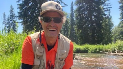 مدرس أمريكي مبتسم يقف بجوار مجرى مائي في منطقة طبيعية، مع أشجار كثيفة في الخلفية، بعد إطلاق سراحه من الاحتجاز في روسيا.