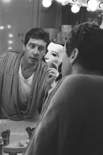 مايكل كروفورد يتأمل في مرآة مع قناع أبيض قبل أداء مسرحي، محاطًا بأضواء مكياج، في خلفية تعكس أجواء برودواي.