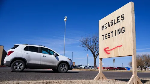 علامة خشبية تشير إلى اختبار الحصبة، مع سيارة تمر بجوارها في منطقة مفتوحة تحت سماء زرقاء.