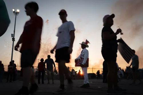 مؤيدون للرئيس ترامب في تجمع حاشد، مع شخص يرتدي زيًا مميزًا يحمل لافتة، بينما تضيء الألعاب النارية في الخلفية.