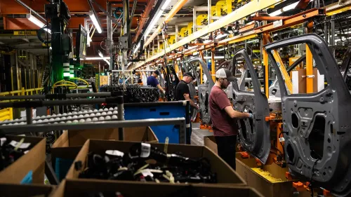 عمال في مصنع يجمعون أجزاء السيارات، مع وجود معدات وأدوات في الخلفية، مما يعكس زيادة في ضغط الأسعار.