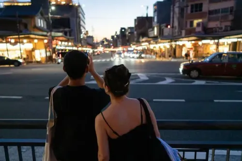 زوجان يقفان في الشارع عند الغسق، يلتقطان صوراً للمدينة المضيئة، مما يعكس زيادة السياحة في اليابان.