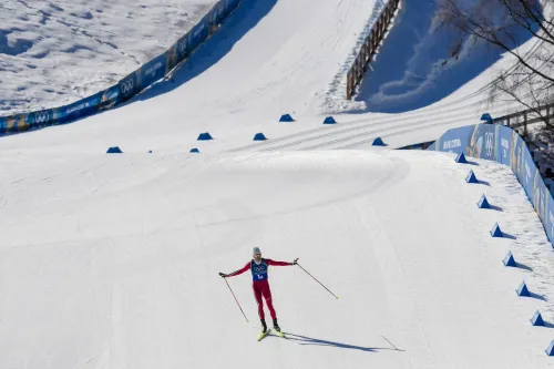 متزلج يرتدي زيًا رياضيًا أحمر، يتزلج على منحدر ثلجي خلال دورة الألعاب الأولمبية الشتوية، مع لافتات تشير إلى المسار في الخلفية.