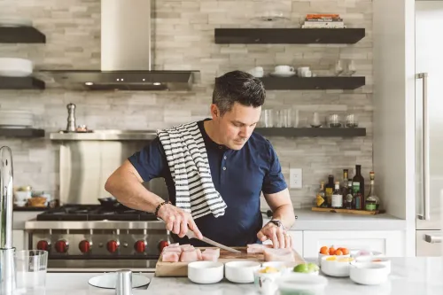 شيف يقف في مطبخ عصري، يقطع الدجاج بينما تحيط به مكونات طهي متنوعة، مستعدًا لإعداد وصفة دجاج وزلابية مستوحاة من جدته.