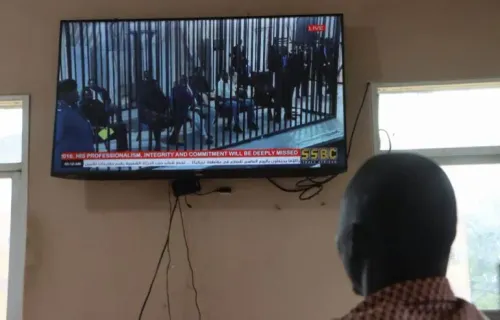 شخص يشاهد بثًا مباشرًا لمحاكمة رياك مشار، النائب الأول لرئيس جنوب السودان، على شاشة تلفاز في قاعة محكمة، مع وجود حواجز أمنية.