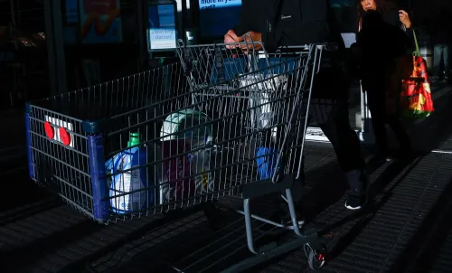 سلة تسوق تحتوي على منتجات متنوعة، تُظهر تأثير التضخم على الأسعار، بينما يمر شخصان بالقرب من المدخل.