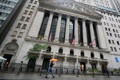 واجهة بورصة نيويورك مع أعمدة ضخمة وأعلام أمريكية، بينما يسير شخص يحمل مظلة برتقالية في يوم ممطر.