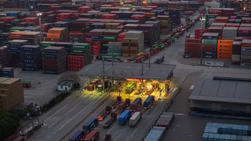 صورة لميناء شحن مزدحم، حيث تتراص حاويات الشحن الملونة في الخلفية، بينما تنتظر الشاحنات في صفوف عند نقطة التفتيش، مما يعكس التوترات التجارية العالمية.