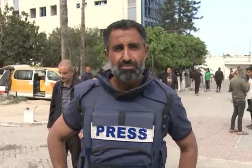 صحفي فلسطيني يرتدي سترة صحافة، يقف في منطقة تعرضت للقصف في غزة، مع وجود أشخاص آخرين في الخلفية، يعكس المخاطر التي يواجهها الإعلاميون.