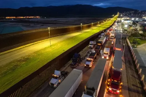 ازدحام مروري لشاحنات النقل على الحدود بين الولايات المتحدة والمكسيك، مع سياج حدودي مضاء، يعكس تأثير الرسوم الجمركية على التجارة.