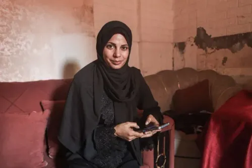 مدلين كلاب، صيادة سمك في غزة، تجلس في غرفة معيشة بسيطة، تحمل هاتفها وتظهر تعابير الأمل والقلق بشأن حصار غزة.