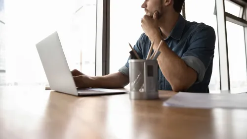 رجل يفكر أثناء استخدامه لجهاز كمبيوتر محمول على مكتب، مع أدوات مكتبية، في سياق الحديث عن تأثير الذكاء الاصطناعي على العمل.