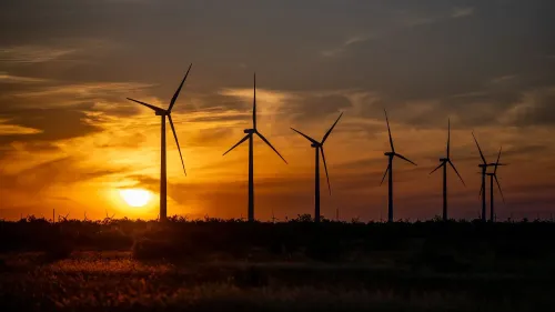 تظهر الصورة مجموعة من توربينات الرياح silhouetted against غروب الشمس، مما يعكس أهمية طاقة الرياح في مواجهة التحديات السياسية.