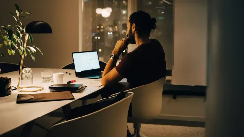 رجل يجلس بمفرده أمام كمبيوتر محمول في مكتب مضاء بشكل خافت، محاط بأدوات العمل، مما يعكس تأثير ساعات العمل الطويلة على الصحة العقلية.