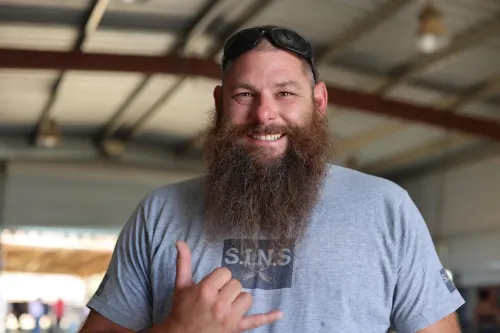 ابتسامة رجل ذو لحية كثيفة يرتدي قميصًا رماديًا، يقف في مكان داخلي، يعكس روح المصارعة والمثابرة بعد حادث مؤسف.