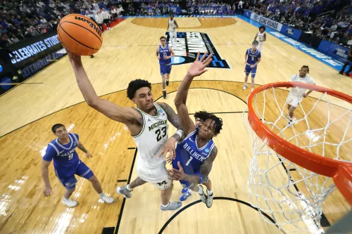 لاعب كرة سلة من فريق ميشيغان يسجل نقطة خلال مباراة NCAA، مع تفاعل قوي من اللاعبين المنافسين في الخلفية.