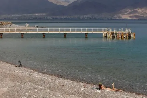 شاطئ مدينة إيلات يظهر رصيفًا خشبيًا يمتد نحو البحر الأحمر، مع شخص مستلقٍ على الرمال. الهجوم بالطائرات المسيرة من الحوثيين يثير القلق.