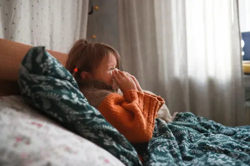 طفلة مريضة تجلس على السرير، تمسك بمنديل وتبدو متعبة، مما يعكس تأثير فيروسات الجهاز التنفسي على الأطفال.