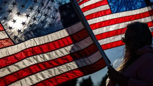 امرأة تحمل علم الولايات المتحدة، مع خلفية مشمسة، تعكس مشاعر الانقسام حول الحلم الأمريكي بين الأجيال المختلفة.