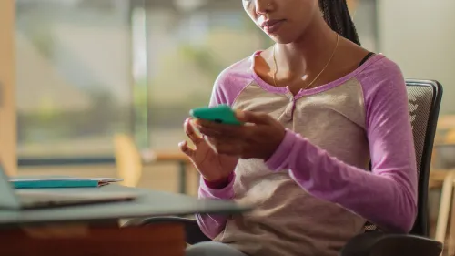 امرأة تجلس أمام الكمبيوتر تمسك بهاتفها الذكي، تبدو مشغولة بالتصفح، مما يعكس تأثير التكنولوجيا على التركيز والإلهاء.