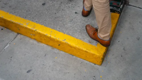 رجل يرتدي حذاءً بنيًا يقف على حافة رصيف أصفر في مدينة مزدحمة، مما يعكس تحديات التنقل في البيئات الحضرية.