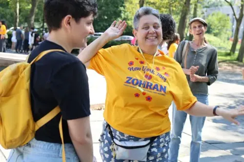 امرأة مبتسمة ترتدي قميصًا أصفر يحمل اسم "زهران" وتشارك في فعالية في نيويورك، محاطة بأشخاص آخرين في أجواء احتفالية.