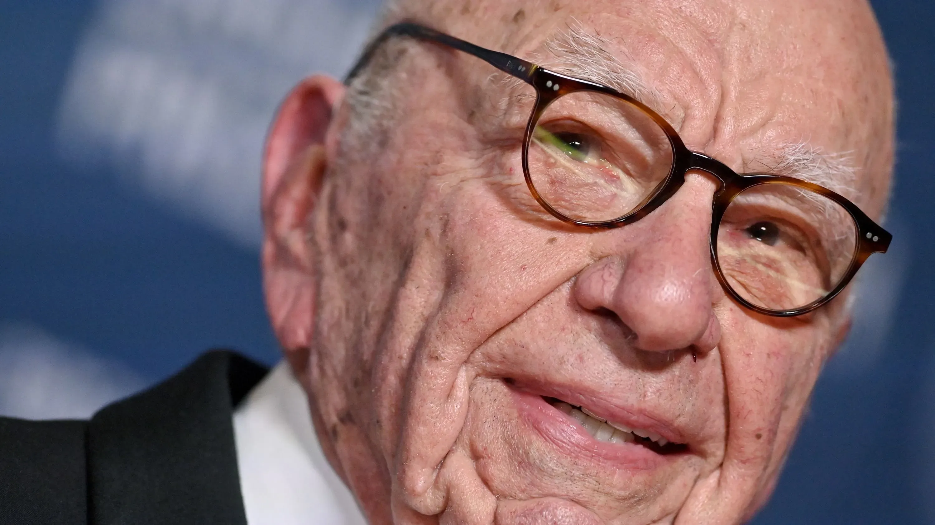 صورة قريبة لرجل مسن يرتدي نظارات، يُعتقد أنه روبرت مردوخ، مع تعبير جدي. تتعلق الصورة بقضية التشهير ضد فوكس نيوز.