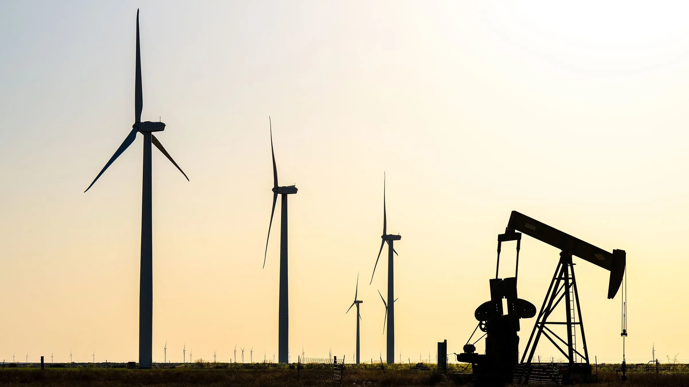 صورة تظهر منصة نفطية في المقدمة مع توربينات رياح في الخلفية، تعكس التحديات التي تواجه صناعة النفط الأمريكية في ظل الضغوط الاقتصادية.