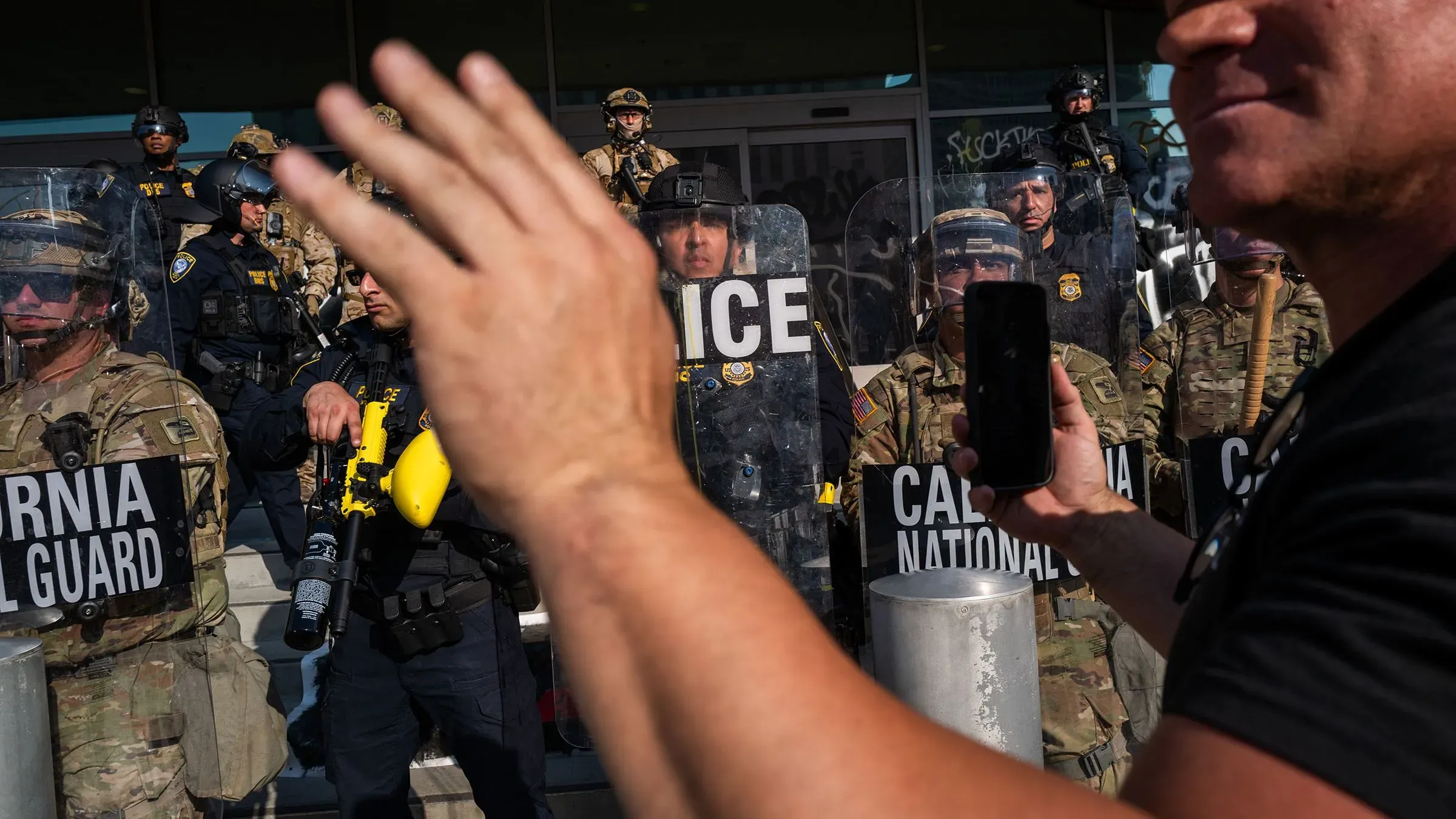 مشهد من احتجاج في لوس أنجلوس يظهر أفراد الحرس الوطني والشرطة، مع شخص يحمل هاتفه لتوثيق الأحداث.