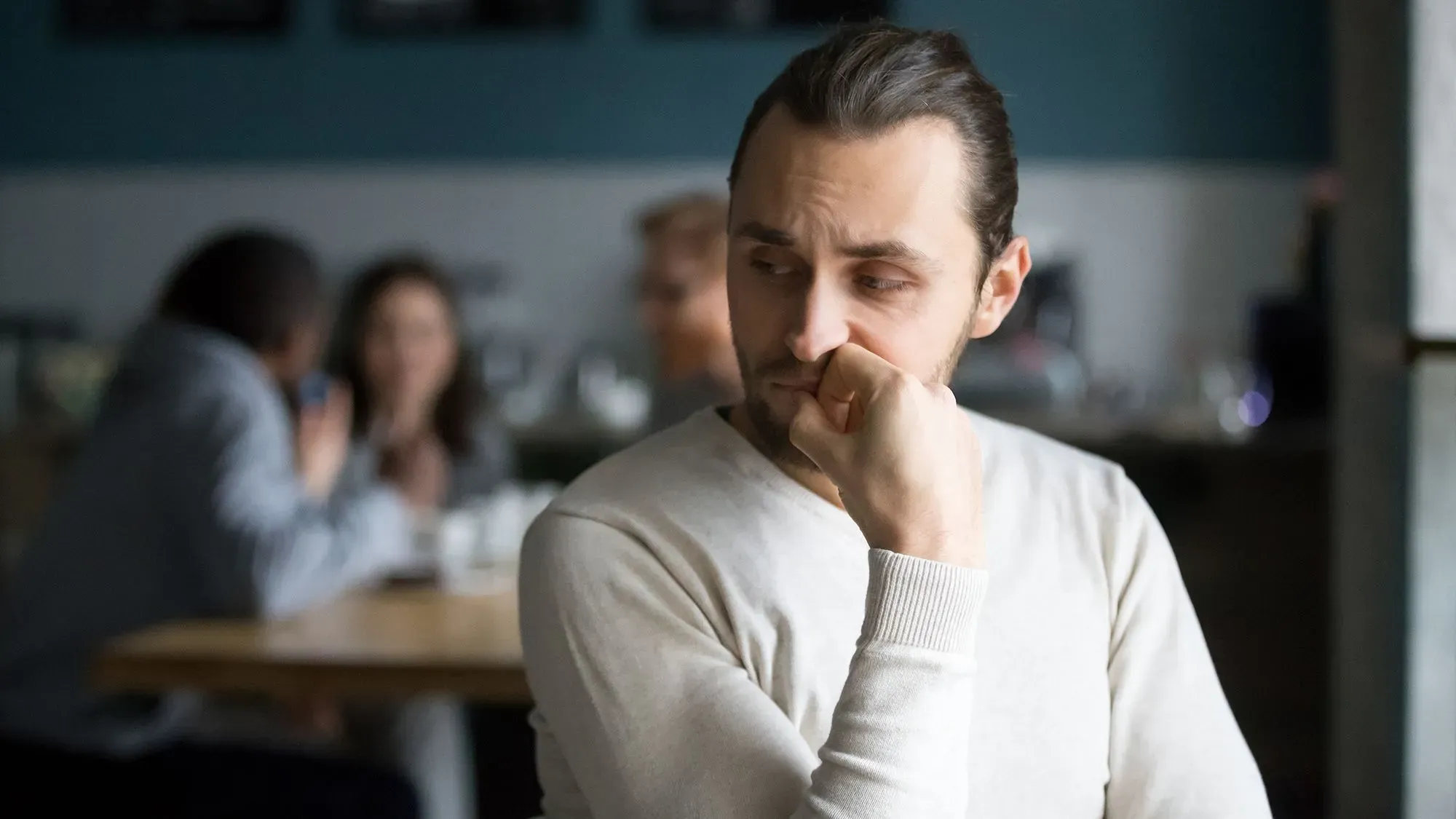 رجل يجلس في مقهى، يبدو عليه التفكير والقلق، بينما يتواجد آخرون في الخلفية يتحدثون معًا.