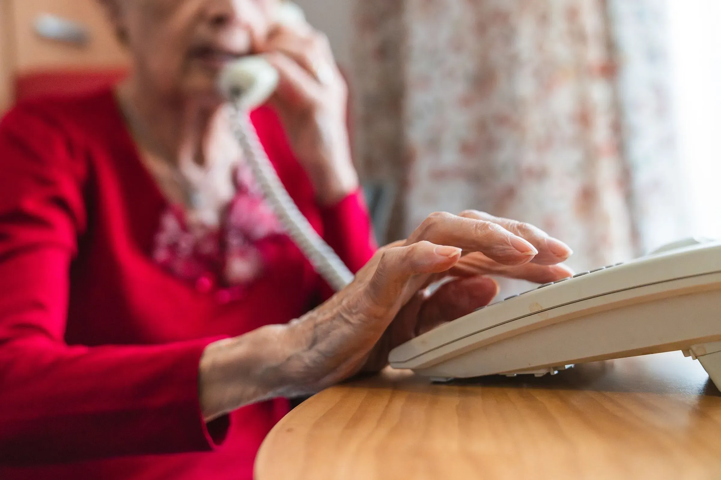 امرأة مسنّة ترتدي قميصًا أحمر وتتحدث في الهاتف الثابت، تعكس الصعوبات التي يواجهها كبار السن في الوصول إلى خدمات الضمان الاجتماعي.