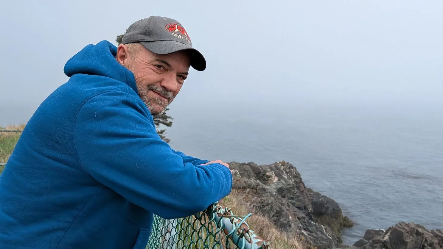 رجل يرتدي سترة زرقاء وقبعة، يستند إلى سياج قرب ساحل ضبابي، مما يعكس الهدوء والطبيعة المحيطة به.